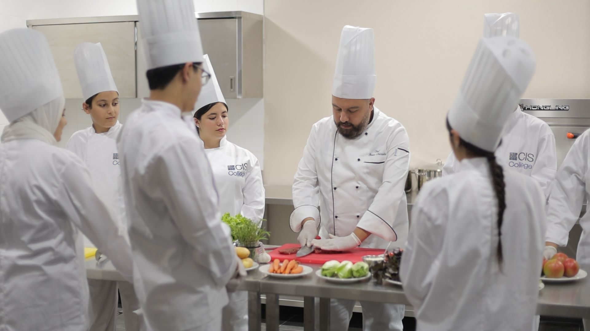 Chef instructor demonstrating food preparation to students in white uniforms in kitchen lab