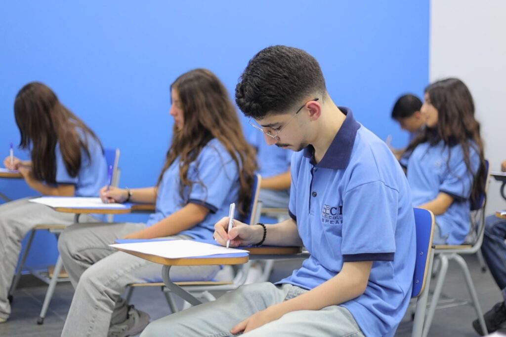 Student in blue uniform writing on paper at classroom desk with classmates in background